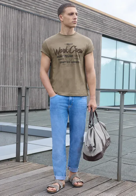 Shirt met ronde hals en Wording Artwork antique beige Een man staat op een dakterras, draagt T-shirt, jeans, sandalen en houdt een tas vast.