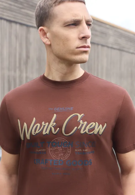 Shirt met ronde hals en Wording Artwork maroon brown Man in een bruin T-shirt met 'Work Crew'-opdruk voor een houten wand.