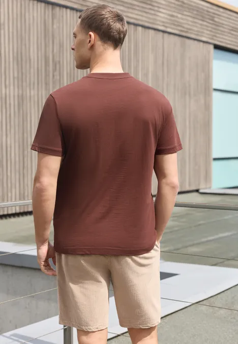 Shirt met ronde hals en Wording Artwork maroon brown Een man in bruin T-shirt en beige korte broek, van achteren voor een modern gebouw.