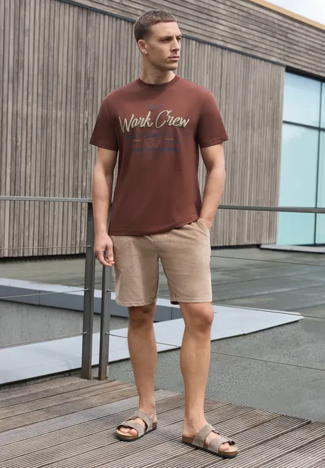 Shirt met ronde hals en Wording Artwork maroon brown Man in bruin T-shirt en beige short op houten terras voor modern gebouw.