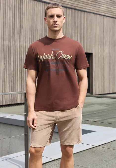 Shirt met ronde hals en Wording Artwork maroon brown Een man in een bruin T-shirt en beige korte broek staat voor een houten gevel.
