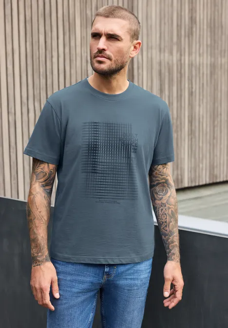 Shirt met ronde hals en opdruk vintage blue Een getatoeëerde man in T-shirt en spijkerbroek staat voor een houten wand en kijkt opzij.