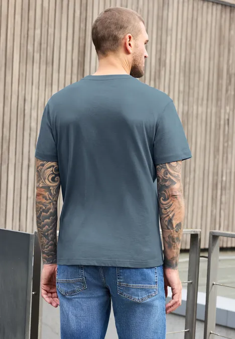 Shirt met ronde hals en opdruk vintage blue Een man met tatoeages op beide armen, in grijs t-shirt en jeans, rug naar de camera voor een houten gevel.