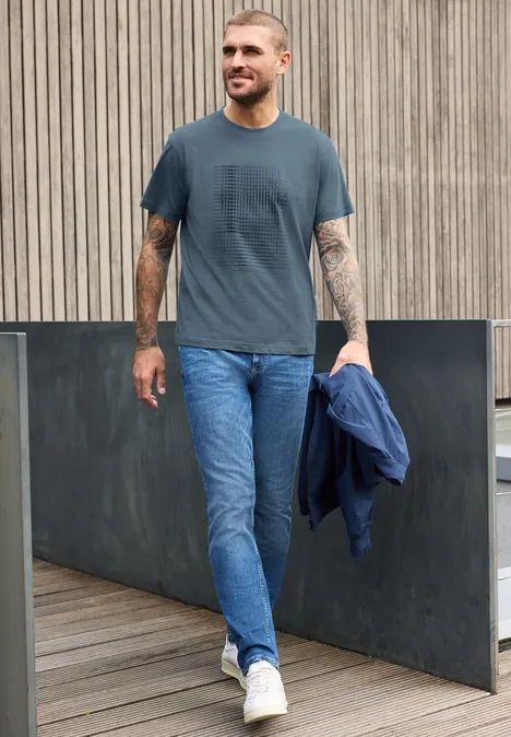 Shirt met ronde hals en opdruk vintage blue Een glimlachende man in jeans en T-shirt draagt een jas en loopt op een houten pad voor een houten wand.