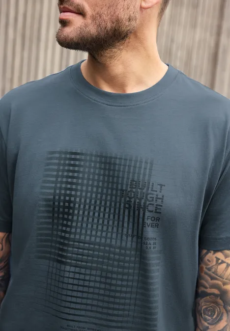 Shirt met ronde hals en opdruk vintage blue Man met baard draagt grijsblauw t-shirt met rasterpatroon en tekst, rozentattoo zichtbaar op arm.