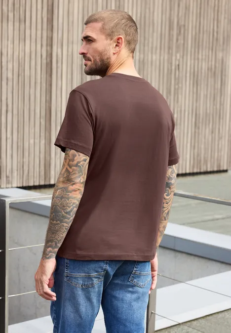 Shirt met ronde hals en print maroon brown Een getatoeëerde man in een bruin T-shirt en spijkerbroek staat buiten voor een houten gevel.