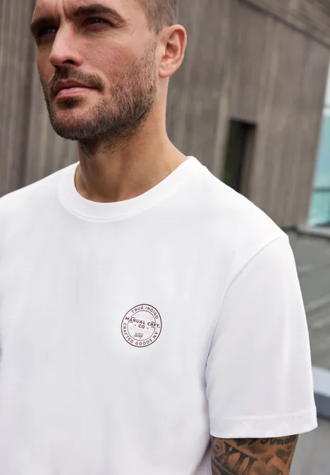Shirt met ronde hals en opdruk op de borst bright white Close-up van een baardige man in wit T-shirt met klein logo, voor een modern gebouw.