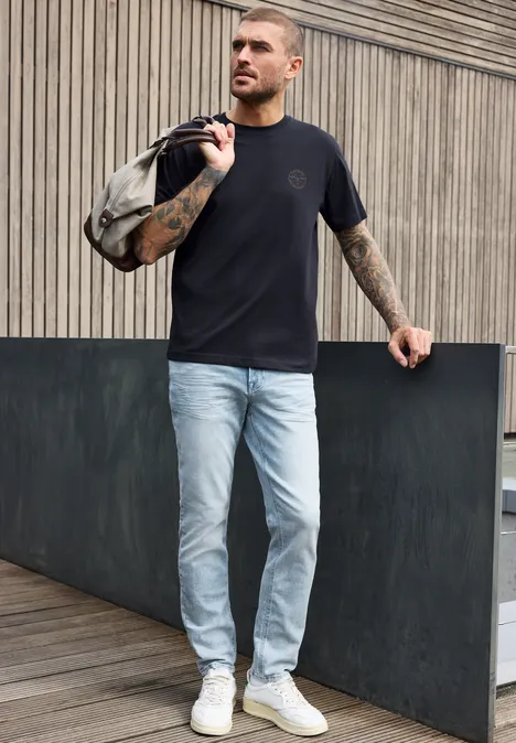 T-Shirt mit Rundhals und Chestprint dark ink blue Ein Mann in schwarzem T-Shirt, hellblauen Jeans und weißen Sneakern trägt eine Tasche über der Schulter vor einer Holzfassade.