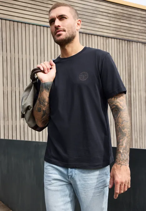 T-Shirt mit Rundhals und Chestprint dark ink blue Ein tätowierter Mann trägt ein schwarzes T-Shirt und hellblaue Jeans, hält lässig einen Rucksack über der Schulter.