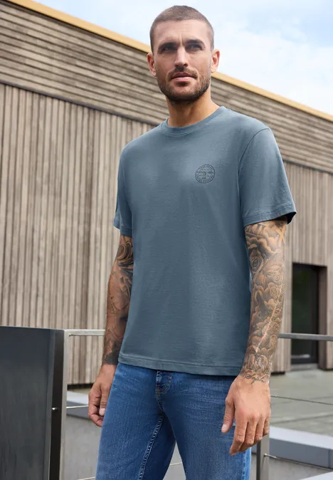 Shirt met ronde hals en opdruk op de borst vintage blue Een man in een blauw T-shirt en spijkerbroek met tatoeages op de armen staat voor een modern houten gebouw.