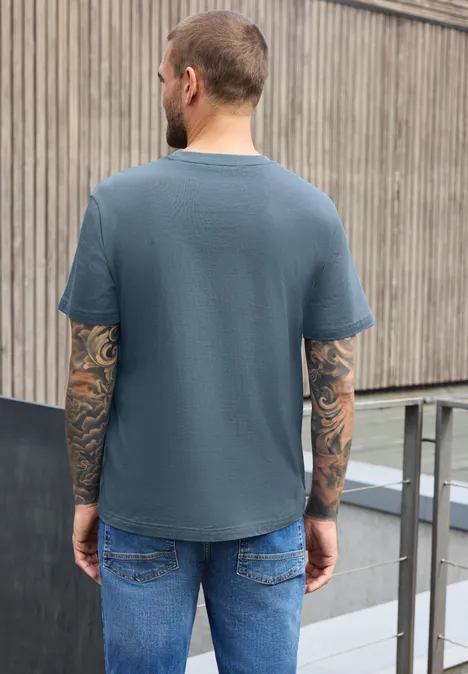 Shirt met ronde hals en opdruk op de borst vintage blue Een getatoeëerde man van achteren in een blauw t-shirt en spijkerbroek leunt tegen een houten hek.