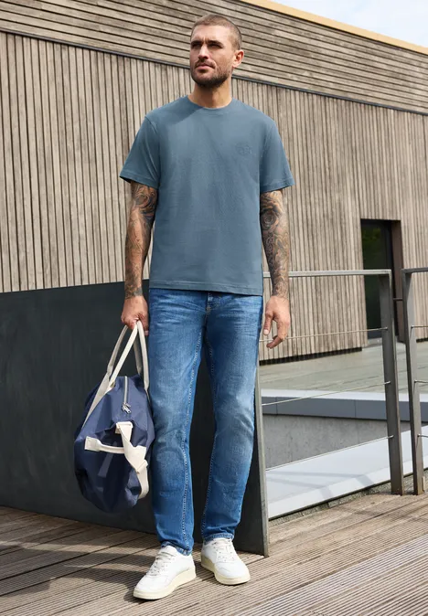 Shirt met ronde hals en opdruk op de borst vintage blue Een man op een houten terras draagt een blauw t-shirt, spijkerbroek, witte sneakers en houdt een blauwe reistas vast.