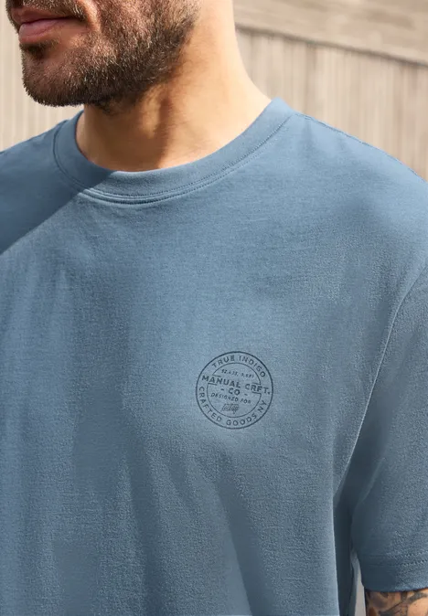 Shirt met ronde hals en opdruk op de borst vintage blue Close-up van een bakkebaarde man in een blauw T-shirt met rond logo op de borst, houtachtergrond.