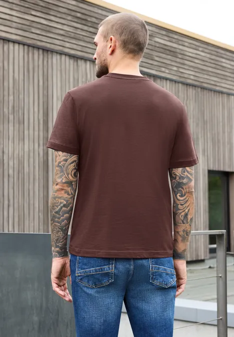 Shirt met ronde hals en opdruk op de borst maroon brown Een man gezien van achteren draagt een bruin t-shirt en spijkerbroek voor een modern houten gebouw.