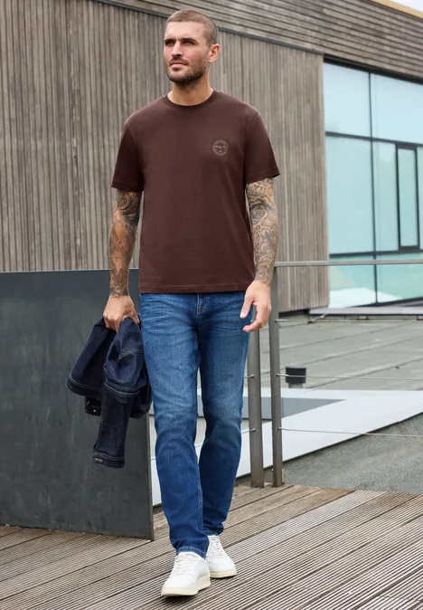 Shirt met ronde hals en opdruk op de borst maroon brown Man in bruin T-shirt, blauwe spijkerbroek en witte sneakers, met jas in hand, loopt over houten vlonder.
