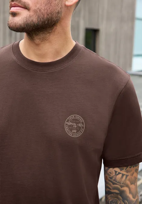 Shirt met ronde hals en opdruk op de borst maroon brown Man draagt bruin t-shirt met klein rond logo op de borst, zichtbare tatoeage op de arm.