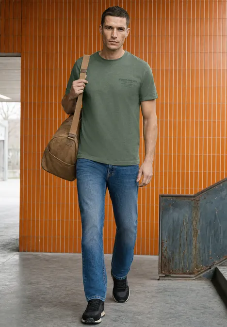 Shirt met ronde hals en opdruk op de borst willow green Man in groen T-shirt en jeans, draagt tas over schouder, voor een oranje muur.