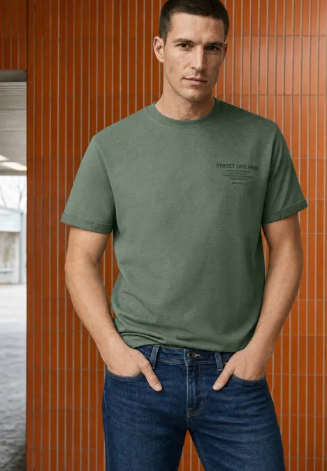 Shirt met ronde hals en opdruk op de borst willow green Een man in een groen T-shirt en jeans staat voor een bruin-oranje wand met verticale panelen.