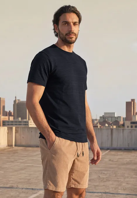 Ein Mann in schwarzem T-Shirt und beigen Shorts steht auf einem Dach und blickt in die Kamera vor Stadtkulisse.