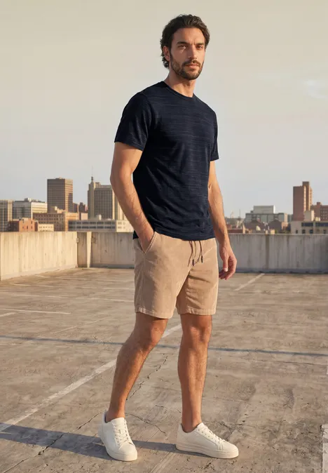 T-Shirt mit Rundhals in Space-Dye-Optik dark ink blue Mann in dunkelblauem T-Shirt, beigen Shorts und weißen Sneakern auf einem Dach mit Stadtkulisse.