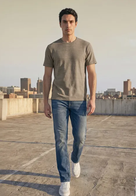 Shirt met ronde hals in space-dye-look antique beige Een man in jeans en t-shirt loopt op een dak met de stad op de achtergrond.