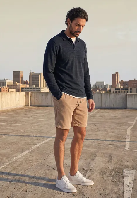 Langarm Poloshirt aus Baumwolle dark ink blue Ein Mann steht auf einem Parkdeck mit Stadtkulisse, trägt dunkelblauen Pullover, beige Shorts und weiße Sneaker.
