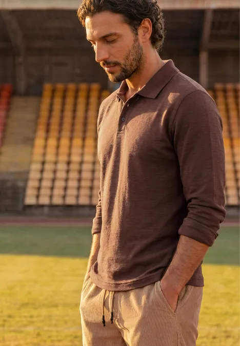 Polo met lange mouwen van katoen maroon brown Een man in een bruin poloshirt met lange mouwen, handen in zijn zakken, staand op het grasveld van een stadion.