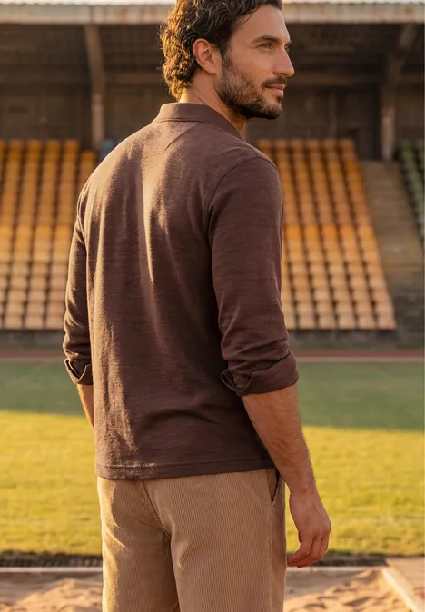 Polo met lange mouwen van katoen maroon brown Een man in bruin poloshirt en beige broek staat op een sportveld voor lege tribunes en kijkt over zijn schouder.