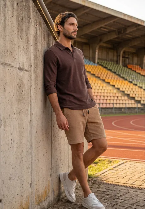 Polo met lange mouwen van katoen maroon brown Een man leunt tegen een betonnen muur op een atletiekbaan, draagt een bruin shirt, beige short en witte sneakers.