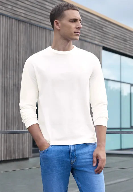 Effen shirt met lange mouwen en ronde hals bright white Een man in een wit langarmshirt en blauwe jeans staat buiten voor een modern gebouw.