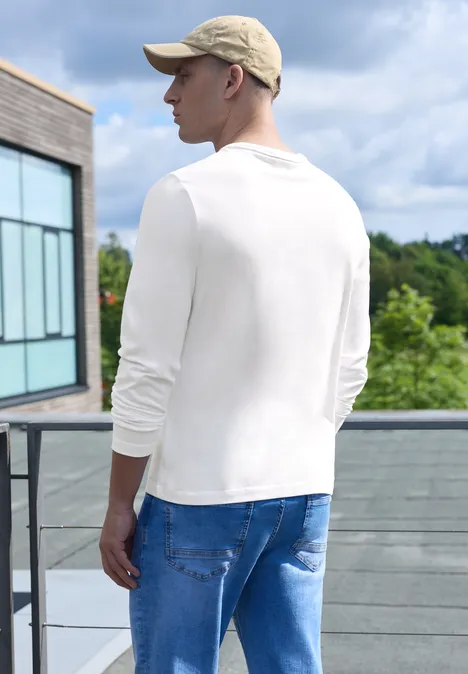 Effen shirt met lange mouwen en ronde hals bright white Een man staat op een terras, draagt een wit langarmshirt, een blauwe spijkerbroek en een pet.