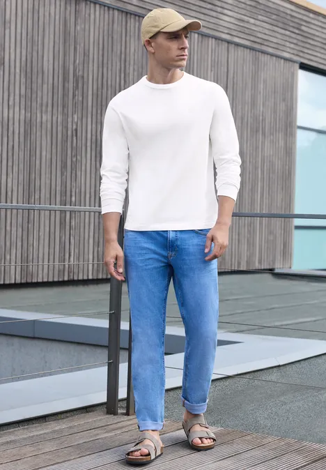 Effen shirt met lange mouwen en ronde hals bright white Een man in jeans, wit shirt en sandalen leunt tegen een reling voor een modern gebouw.
