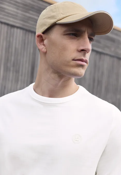 Effen shirt met lange mouwen en ronde hals bright white Een man met een beige pet en wit T-shirt voor een houten muur