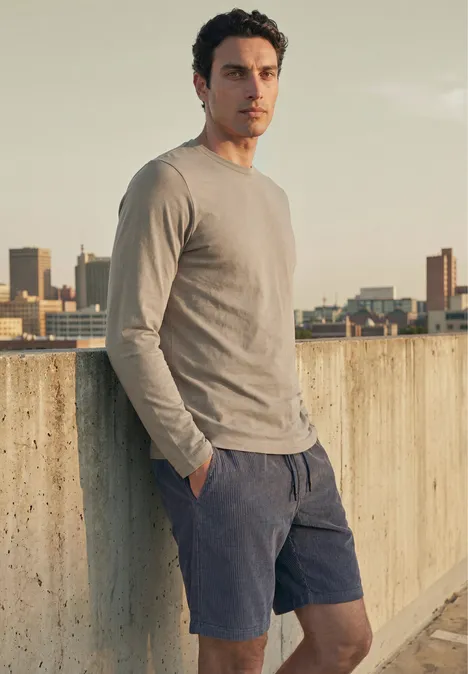Effen shirt met lange mouwen en ronde hals antique beige Een man leunt nonchalant tegen een betonnen muur in de stad, draagt een longsleeve en shorts, kijkt naar de camera.
