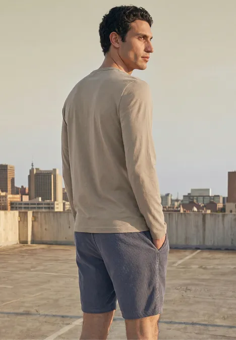 Effen shirt met lange mouwen en ronde hals antique beige Een man staat op een dak en kijkt over de stad, draagt een beige longsleeve en blauwe short.
