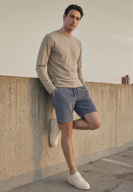 Effen shirt met lange mouwen en ronde hals antique beige Jonge man in beige trui en blauwe short leunt ontspannen tegen een betonnen muur op een dak.