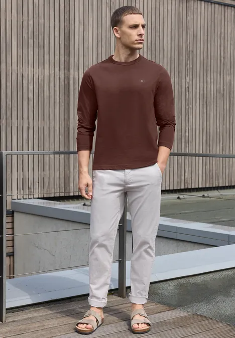 Effen shirt met lange mouwen en ronde hals maroon brown Een man staat voor een houten gevel, draagt een bruine trui, lichte chino’s en sandalen.