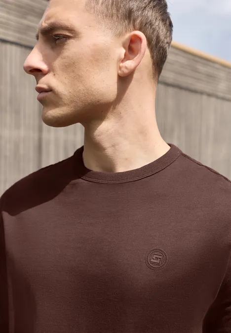 Effen shirt met lange mouwen en ronde hals maroon brown Profielfoto van een man in een bruin T-shirt voor een houten gevel.