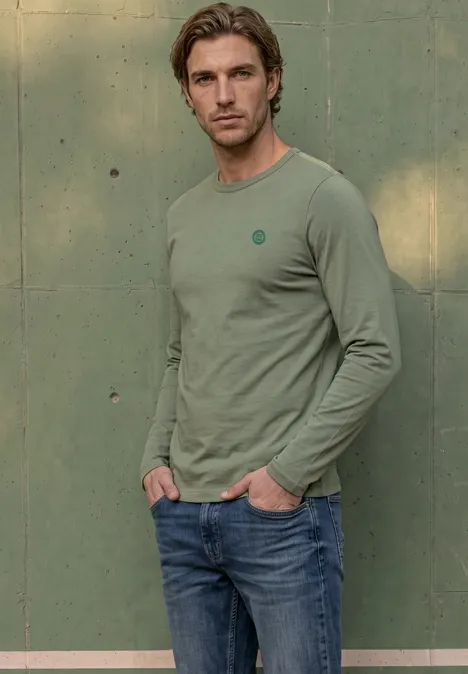 Ein junger Mann mit hellbraunen Haaren trägt ein grünes Langarmshirt und Jeans vor einer grünen Wand.