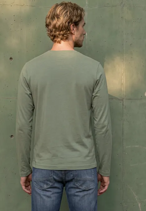 Ein Mann von hinten in legerem, grünen Langarmshirt und Jeans steht vor einer grünen Wand.