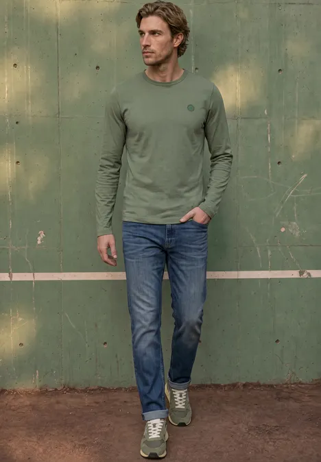 Ein Mann in grünem Langarmshirt, Jeans und Sneakers posiert vor einer grünen Wand auf einem Tennisplatz.