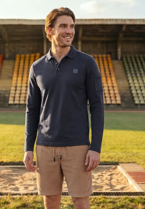 Langarm Poloshirt mit Taschendetail dark ink blue Mann in navyblauem Langarm-Poloshirt und beigen Shorts lächelt auf einer Leichtathletikbahn vor leeren Tribünen.