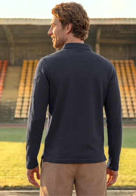 Langarm Poloshirt mit Taschendetail dark ink blue Ein Mann steht mit dem Rücken zur Kamera auf einem Stadionrasen, trägt blaues Langarmshirt und braune Hose.