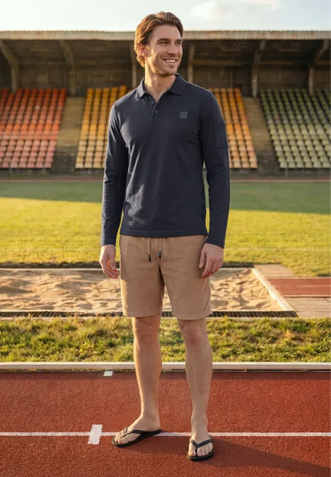 Langarm Poloshirt mit Taschendetail dark ink blue Ein Mann steht entspannt auf der Laufbahn vor bunten Tribünen, trägt Poloshirt, Cargo-Shorts und Flip-Flops.