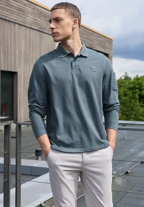 Poloshirt met lange mouwen en zakdetail vintage blue Een man staat op een balkon, draagt een groen poloshirt met lange mouwen en een lichte broek, kijkt opzij.