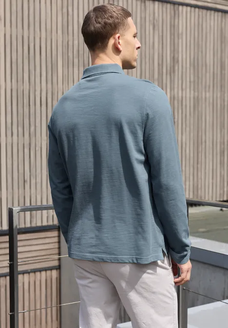 Poloshirt met lange mouwen en zakdetail vintage blue Een man van achteren draagt een petrolgroene lange-mouwen polo en lichte broek op een balkon.
