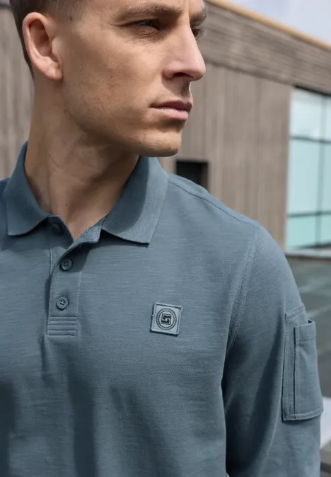 Poloshirt met lange mouwen en zakdetail vintage blue Een man in profiel draagt een grijsblauw poloshirt met borstpatch en armenzak voor een modern gebouw.