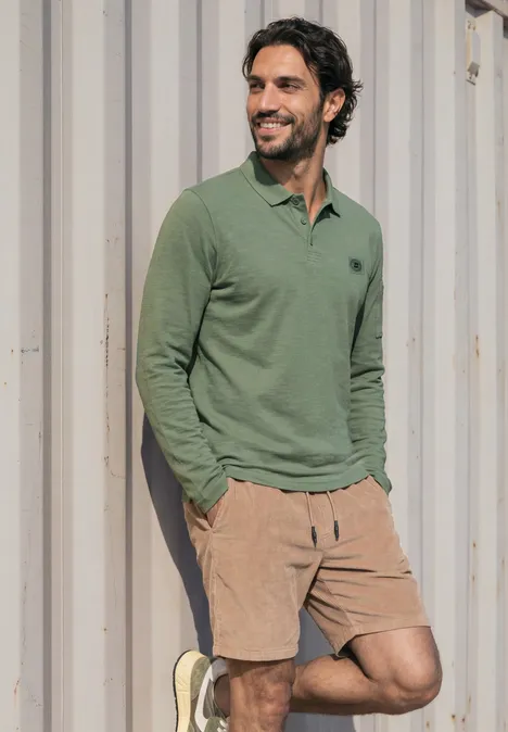 Poloshirt met lange mouwen en zakdetail willow green Man leunt lachend, draagt groen poloshirt en beige korte broek bij metalen container.