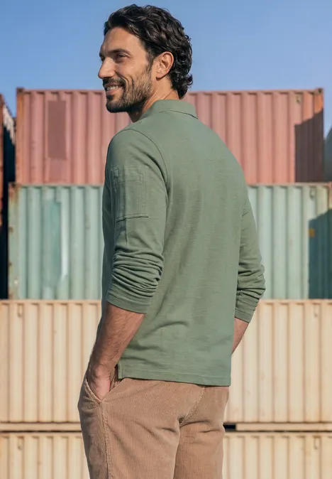 Poloshirt met lange mouwen en zakdetail willow green Een baardige man in groen shirt en bruine broek kijkt over zijn schouder bij kleurrijke containeropslag.