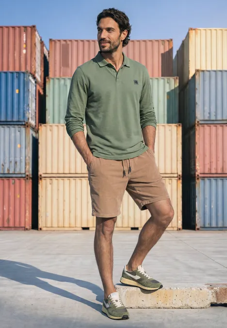 Poloshirt met lange mouwen en zakdetail willow green Een man in een groen poloshirt, bruine shorts en groene sneakers staat voor gekleurde containers.